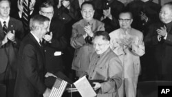 1979年8月28日,美国副总统蒙代尔和中国副总理邓小平在两国文化和水电协议书签署后,交换协议文本。前排鼓掌者有中国总理华国锋(中)、美国驻华大使伦纳德·伍德科克(华国锋左侧)和中国外交部长黄华(华国锋右侧)